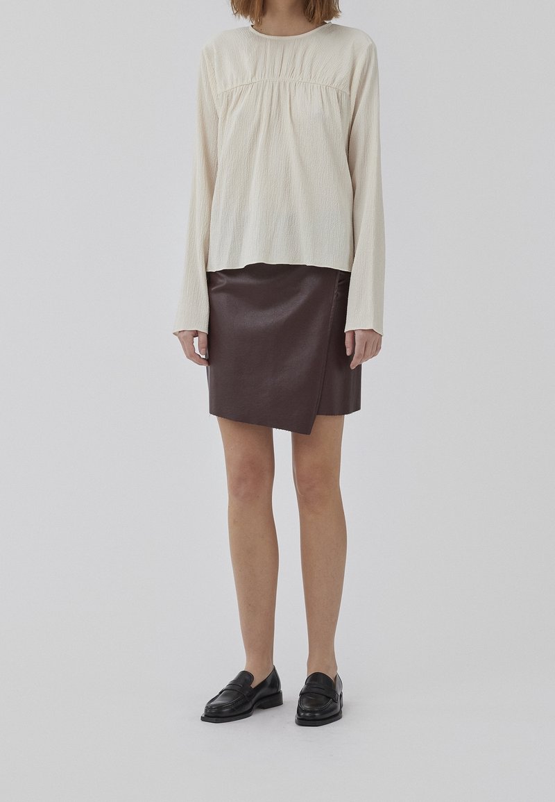 Maglia a maniche lunghe di colore crema texture, abbinata a una gonna a portafoglio in finta pelle marrone. Mocassini neri completano l'outfit. Design semplice e moderno.