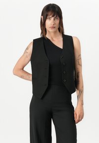 Svart skreddersydd vest med fem knapper, med en tettsittende design, uten ermer, og subtile sidelommer, båret over en svart topp.