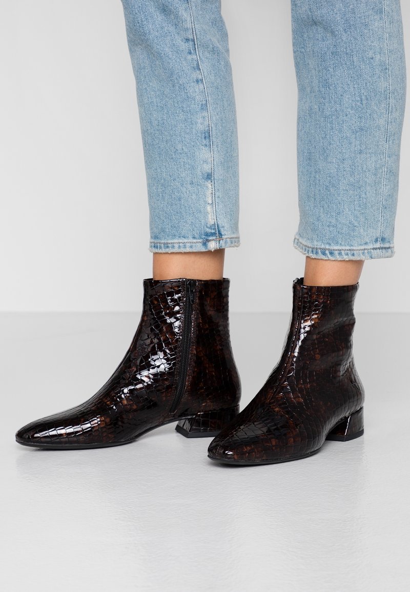 Personne portant un jean court bleu clair et des bottines à motif croco brun foncé brillant avec des talons blocs bas et des fermetures éclair latérales.