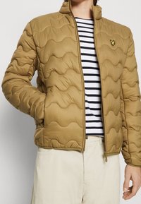 Persona que lleva una chaqueta acolchada color beige con un logo de pájaro amarillo, una camiseta de rayas blancas y negras, y pantalones color off-white, con una mano en el bolsillo de la chaqueta.