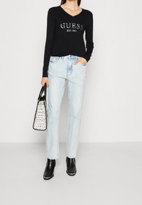 Zwarte lange mouw top met zilveren "GUESS" logo, lichtblauwe jeans met gescheurde details, zwarte enkellaarsjes en een handtas met patroon.