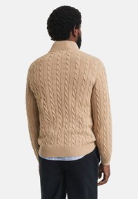 Beige Kabelstrickpullover mit hohem Kragen, geripptem Saum und strukturiertem Muster, ausgestattet mit langen Ärmeln und einer taillierten Silhouette.