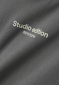 Grått stoff med trykt tekst "Studio edition" og koordinater "55°57'03''N." Glatt tekstur med en subtil ribbet mønster.