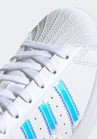 Vit sneaker med texturerad ovandel, featuring iriserande blå accenter och klassisk tre-stripes design. Platta snören och perforerad detaljering.