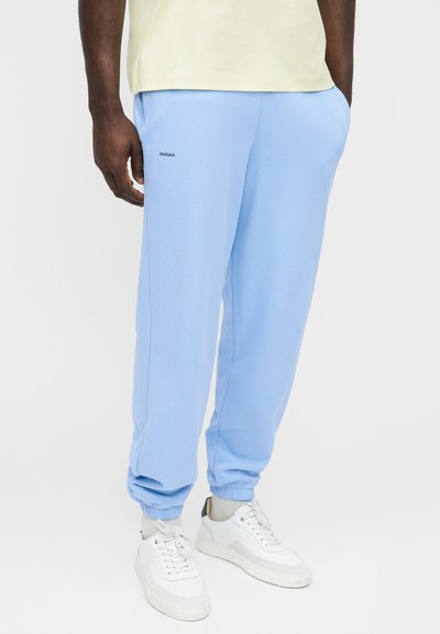 Férfi világoskék, gumis mandzsettás melegítőnadrágban és fehér tornacipőben, egyik kezét a zsebében tartva áll.