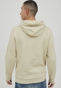 Felpa beige con finitura testurizzata, dotata di cappuccio con cordino, polsini e orlo a costine, indossata con jeans blu. Vista da dietro.