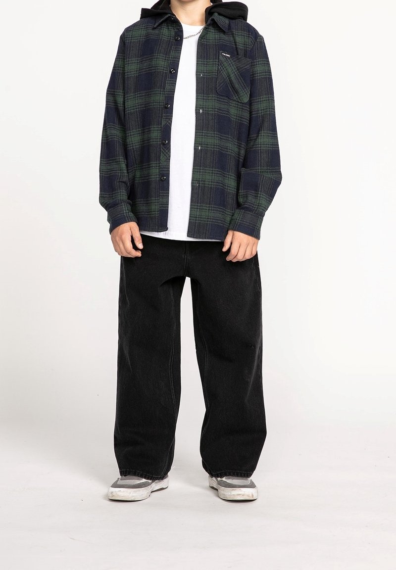 Personne portant une chemise blanche sous une chemise à carreaux vert foncé et noir avec boutons, un pantalon large noir et des baskets grises.