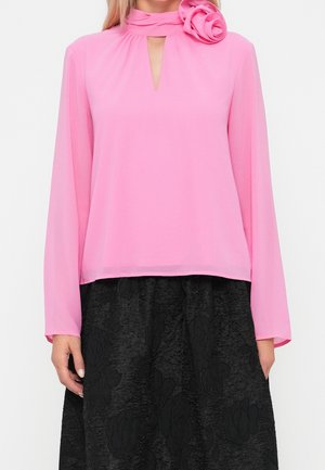 Long-sleeve pink blouse with keyhole neckline and rose detail at collar paired with textured black skirt.