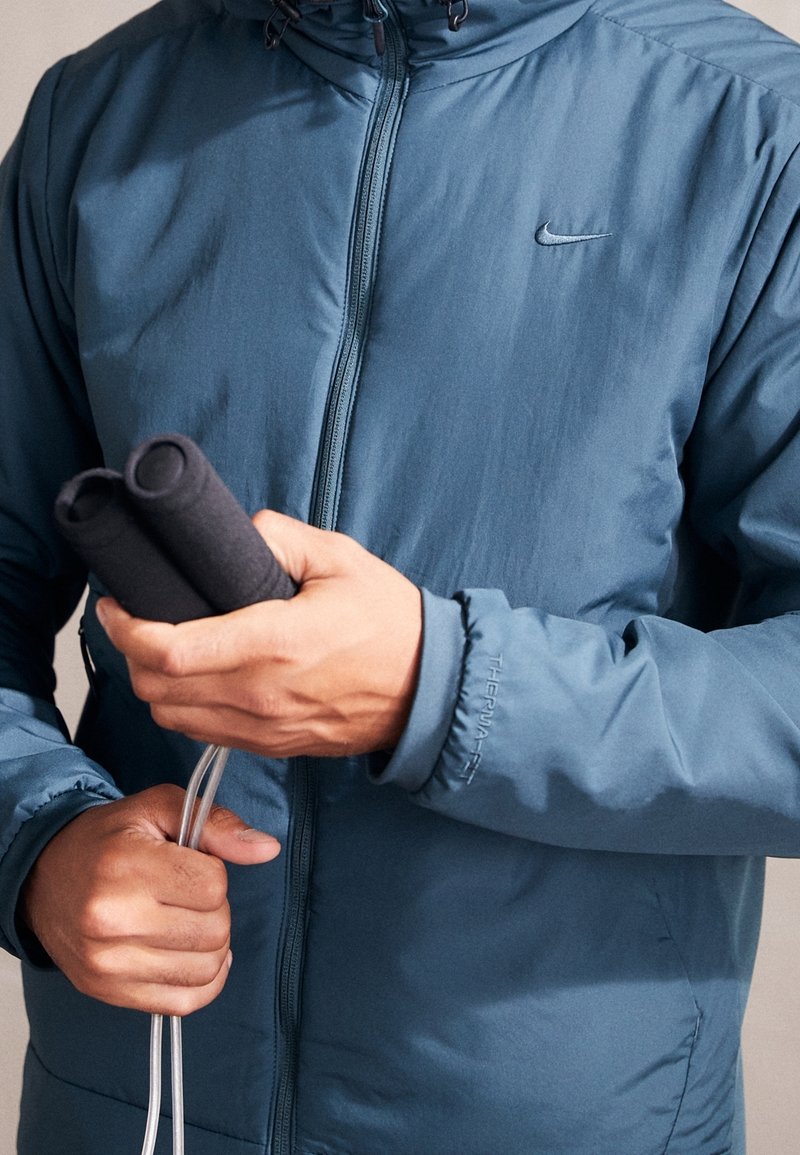 Personne portant une veste Nike Therma-FIT bleue tenant une corde à sauter avec une poignée noire des deux mains.