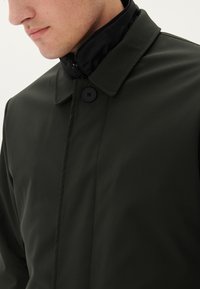 Abrigo impermeable verde con alto cuello, que presenta un cierre frontal con botones y una textura suave. Incluye un forro de cuello negro en contraste.