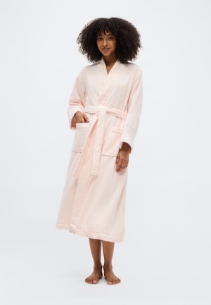 Ženska s kodrastimi lasmi, bosa, oblečena v dolgo bledo rožnato kopalno haljo z žepi in pasom, stoji na belem ozadju.