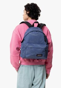 Sac à dos Eastpak bleu en tissu texturé, avec une grande poche avant, des bretelles noires et un logo. Porté par-dessus un sweat-shirt rose.