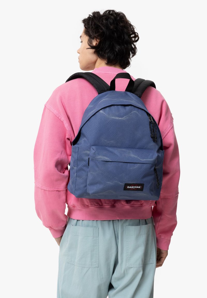 Sac à dos Eastpak bleu en tissu texturé, avec une grande poche avant, des bretelles noires et un logo. Porté par-dessus un sweat-shirt rose.