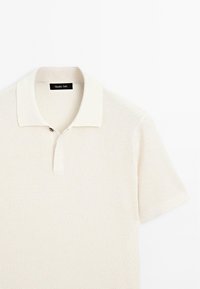 Polo a maniche corte color crema, caratterizzato da un design a maglia testurizzata, colletto con bottoni e etichetta dettagliata sul collo.