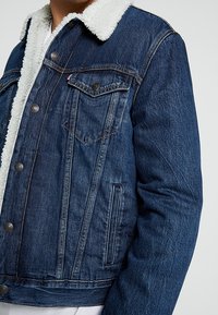 Jeansjacka med krage i fårskinnstyg, mörkblå färg, två bröstfickor och sidosfickor, med metallknappar och synliga sömdetaljer.