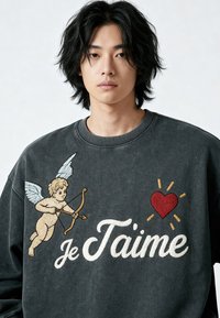 Jeune homme portant un sweat-shirt sombre avec un chérubin brodé, un cœur rouge et le texte "Je T'aime" sur le devant, sur un fond uni.