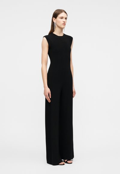 Γυναίκα που στέκεται, φοράει μαύρη ολόσωμη φόρμα χωρίς μανίκια με φαρδιά πόδια και μακριά, κρεμαστά σκουλαρίκια, σε απλό ανοιχτό φόντο.