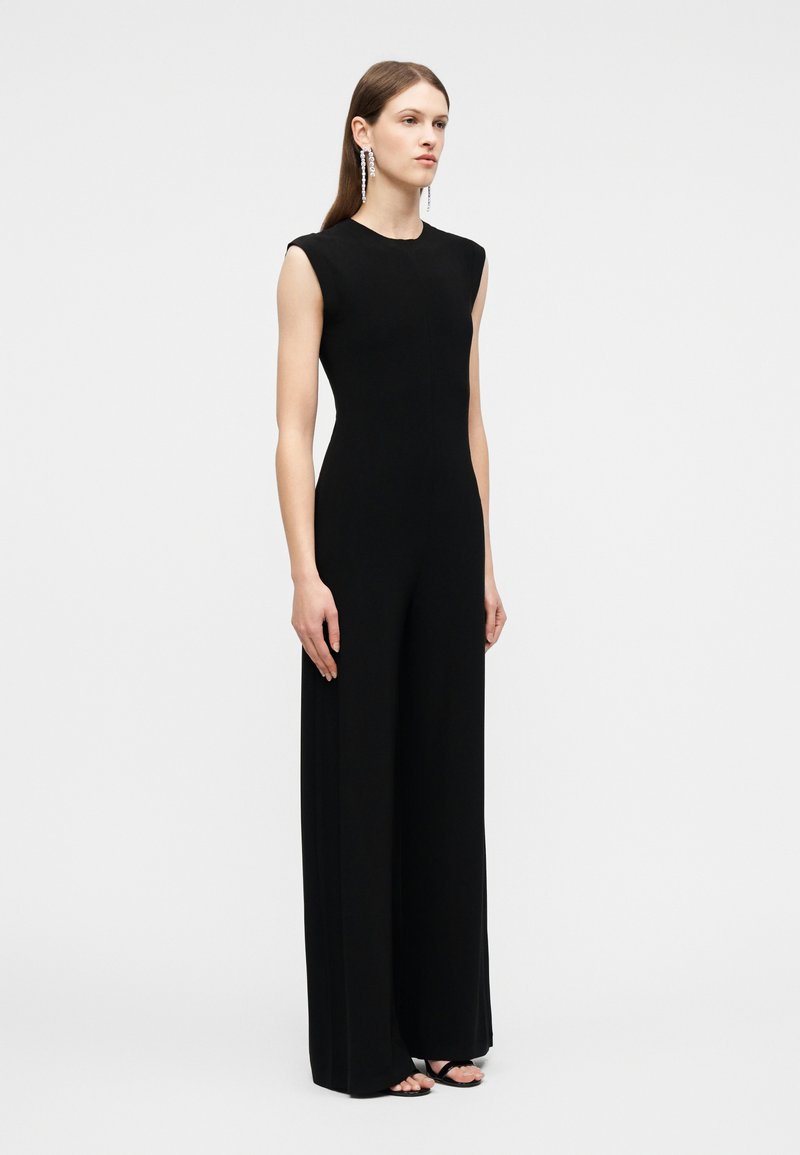 Femme debout, portant une combinaison noire sans manches à jambes larges et de longues boucles d'oreilles pendantes, sur un fond clair uni.