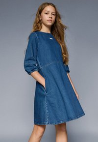 Denim obleka s tremi četrtinami rokavi, okroglo izrezom, stranskimi žepi in obrabljenim robom. V srednjem modrem odtenku z diskretnimi šivi.