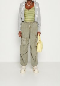 Felpa grigia corta con testo bianco, canotta a righe verdi, pantaloni cargo oliva con tasche, borsa gialla e scarpe con plateau beige.