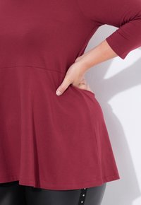 Burgunderfarbenes Oberteil mit Dreiviertelärmeln und ausgestellt geschnittenem Saum, aus weichem Stoff. Die Hand des Models ruht auf der Hüfte und zeigt die Seitentaschen.