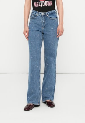 Mujer vestida con jeans de tiro alto azul con pequeños bordados florales blancos, una camiseta gráfica negra y bailarinas rojo oscuro, de pie contra un fondo blanco.