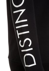 Svart långärmad topp med djärv vit text som läser "DISTINCT" längs vänster ärm. Materialet verkar mjukt med en jämn textur.