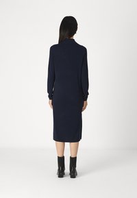 Abito a maglia blu navy con maniche lunghe e spacco laterale, caratterizzato da un design aderente e una silhouette dritta, abbinato a stivali neri con tacco.
