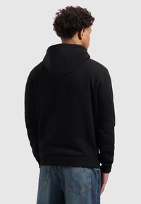 Man met krullend haar in een zwarte hoodie en blauwe spijkerbroek, met de rug naar de camera gekeerd tegen een effen lichte achtergrond.