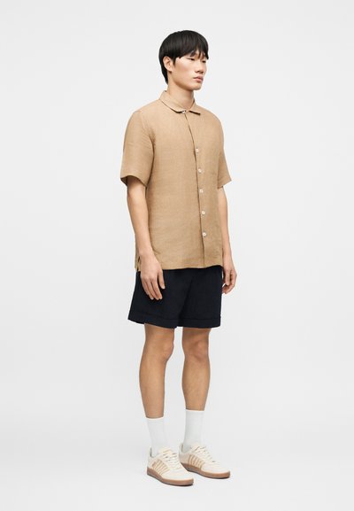 Κοντομάνικο, ανοιχτό καφέ λινό πουκάμισο με διπλωμένο γιακά, λευκά κουμπιά, σετ με σκούρα σορτς και μπεζ αθλητικά παπούτσια. Μινιμαλιστικός σχεδιασμός.