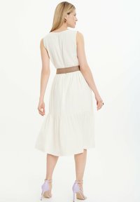 Vestido sin mangas color beige claro con un dobladillo en capas, que presenta un cinturón marrón texturizado. La modelo lleva tacones lila, y el fondo es blanco.