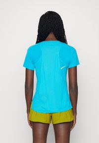Tyrkysové sportovní tričko s krátkými rukávy, hladkou texturou a plochými švy. Na zadní straně má bílé logo; v kombinaci se zelenými šortkami.