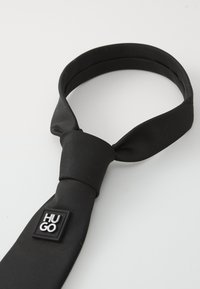 Papion negru din țesătură netedă cu textură țesută. Prezintă un design legat și un etichetă pătrată cu "HUGO" în negru și argintiu.
