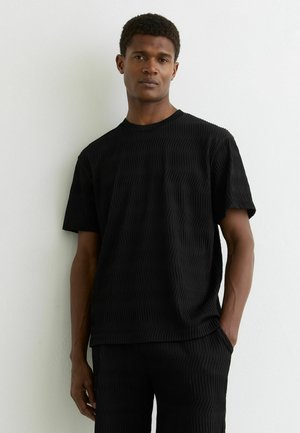 Férfi áll fehér fal előtt, fekete mintás rövid ujjú ingben és hozzáillő nadrágban, egyik kezét a zsebében tartva.