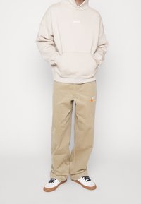 Personne portant un sweat à capuche beige avec poche kangourou, un pantalon large en velours côtelé couleur beige avec le logo Nike, et des baskets blanches avec semelles en gomme et lacets noirs.
