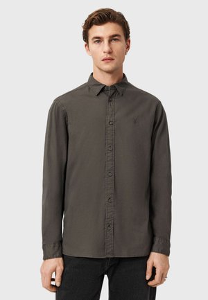 Junger Mann mit kurzem braunem Haar, der ein dunkelgraues Hemd mit langen Ärmeln und schwarzen Hosen vor einem schlichten Hintergrund trägt.