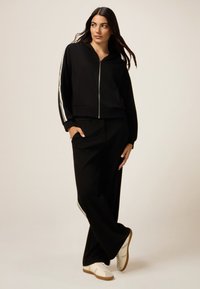 Felpa nera con zip e strisce laterali bianche, abbinata a pantaloni neri coordinati. Presenta una vestibilità comoda e sneakers bianche con suole in gomma.