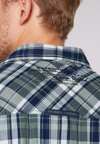 Close-up van de bovenrug van een man die een groen-blauw geruit overhemd draagt met geborduurde tekst en een "Green Label" tag op de kraag.