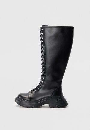 Vysoká čierna kožená šnurovacia čižma s masívnou podrážkou a striebornými očkami na jednoduchom svetlom pozadí.