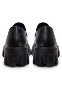 Chaussures à plateforme noires et épaisses vues de l'arrière, avec des tiges en cuir lisse et des semelles à profondes rainures sur un fond blanc.