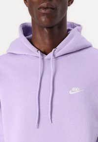 Violetti Nike-huppari pehmeästä kankaasta, jossa on kiristysnauhalla varustettu huppu ja pieni valkoinen logo vasemmassa rinnassa.