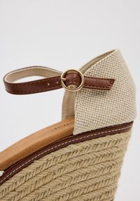 Sandalia de cuña con parte superior de tela beige trenzada y tira de cuero marrón, con hebilla dorada. Plataforma de yute texturizada.