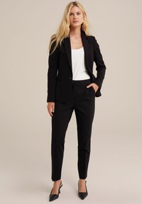 Černý blazer a kalhoty na míru, hladký materiál, úzký střih, přední kapsy, v kombinaci s bílým topem a černými podpatky. Jednoduchý design.