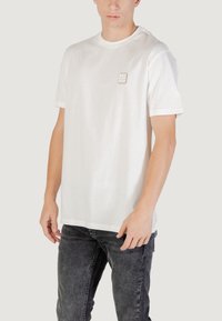 T-shirt in cotone bianco con collo a giro, maniche corte e un piccolo logo sul petto sinistro; abbinato a jeans grigio scuro.