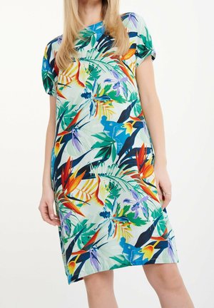 Frau trägt ein kurzärmeliges, knielanges Kleid mit einem bunten tropischen Blumenmuster auf hellem Hintergrund.