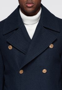 Marineblå dobbeltradet uldblandet frakke med store gyldne knapper og brede revers, lag på lag over en hvid strikket rullekrave.