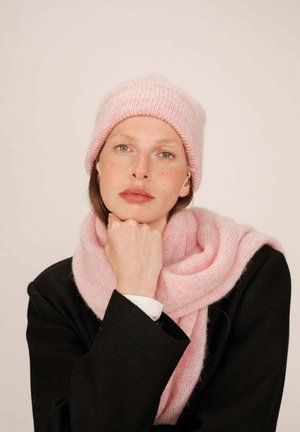 Un bonnet en tricot rose clair et une écharpe en matériau doux. Le modèle porte un blazer noir avec une chemise blanche en dessous, posant avec le menton reposant sur la main.