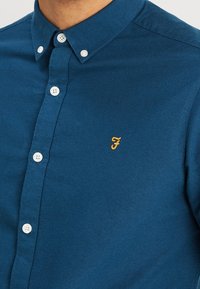 Niebieska koszula z guzikami w gładkim materiale; posiada białe guziki i haftowane żółte logo "F" na klatce piersiowej.