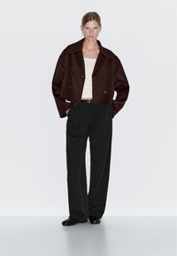 Brązowa krótka marynarka dwurzędowa z czarnymi guzikami, zestawiona z kremowym topem na ramiączkach i luźnymi czarnymi spodniami. Czarne buty dopełniają całość.