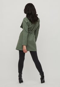 Abrigo envolvente de color verde oliva con cintura ceñida, mangas largas y una textura suave, combinado con jeans skinny negros y botas de tobillo.